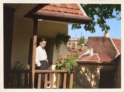 A color photograph of a man on a sunlit balcony, with red rooftops and blue sky